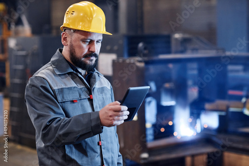Confident male engineer or builder in workwear using digital tablet for checking data about goods ordered by online clients