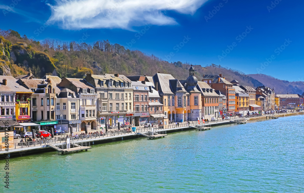 Dinant, Belgium - March 9. 2022: View over river meuse on waterfront promenade with ancient colorful houses against blue winter sky, fluffy clouds