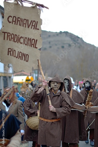 Enciso, Spain – March 5, 2022: Disguised man with brown cloth and tree bark mask acting as a standard bearer followed by a retinue of disguised people. Traditional carnival of Enciso.