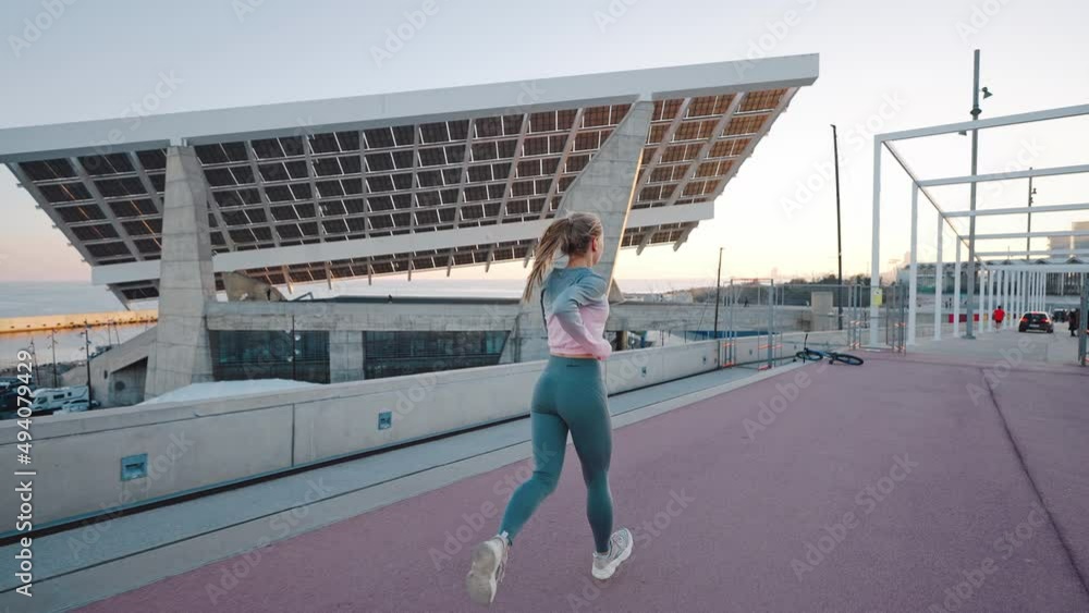 Camera follow from back energetic active young female jogger running fast by wide bridge in urban area. Seashore and big city glass and metal constructions are on background. Rear view. Slow motion Camera follow from back energetic active young female jogger running fast by wide bridge in urban area. Seashore and big city glass and metal constructions are on background. Rear view. Slow motion