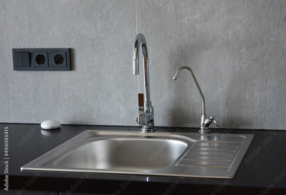 A metal kitchen sink on a black kitchen countertop with two water faucets, where one is a small filter faucet for drinking water. A metal kitchen sink on a black kitchen countertop with two water faucets, where one is a small filter faucet for drinking water.