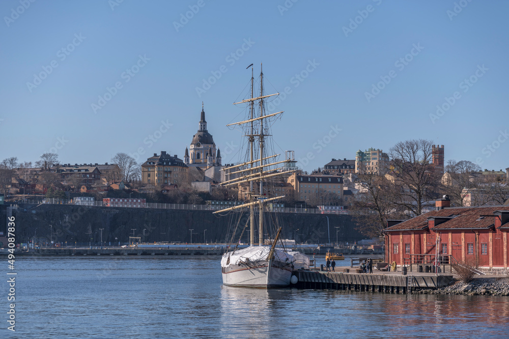 Fototapeta premium Sail boat, the brig Tre Kronor af Stockholm at a jetty on the island Kastellholmen and the district Södermalm with church and towers a sunny spring day in Stockholm