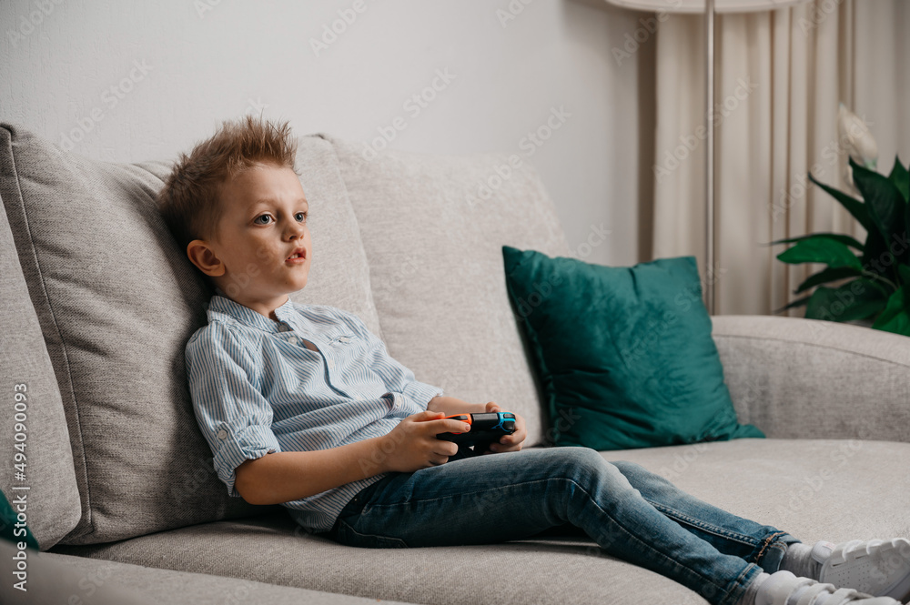 Happy boy playing video games holding game controller sitting on the coach in living room Happy boy playing video games holding game controller sitting on the coach in living room