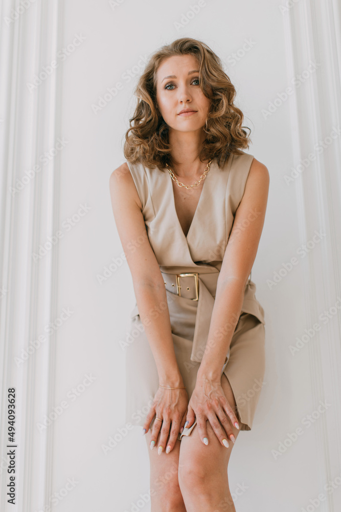 Bottom-up fashion portrait of young woman dressed in a short formal dress stands against a white wall with moldings and looks into the camera Bottom-up fashion portrait of young woman dressed in a short formal dress stands against a white wall with moldings and looks into the camera