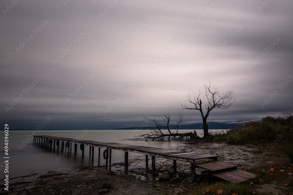 View of a pier on a lake, beneath a dramatic, moody sky with skeletal, bare trees on the background