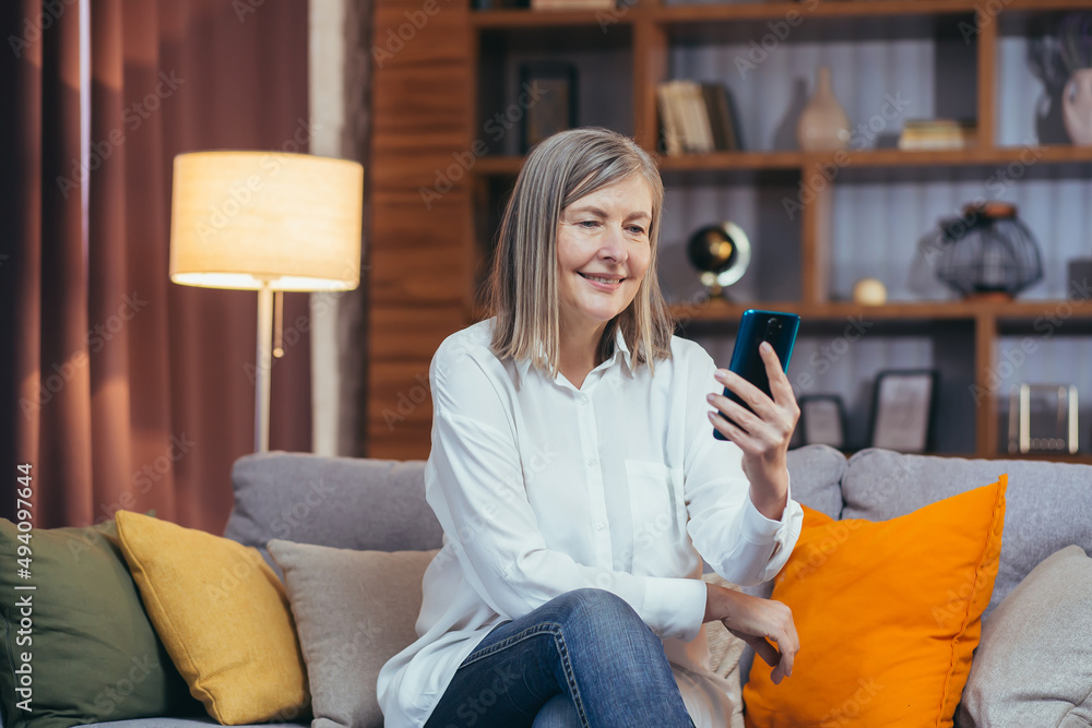 happy senior woman uses phone for online communication, sitting on sofa at home in the evening and smiling happy senior woman uses phone for online communication, sitting on sofa at home in the evening and smiling