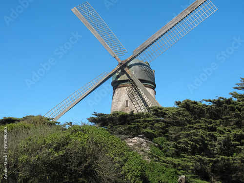 2009 Dutch Windmill at Golden Gate Park sits atop textured greenery covered hillside against bright blue sky with blades crosswise.