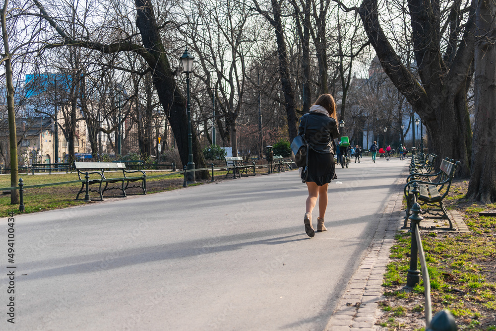A lonely girl in a short leather jacket and skirt with a bag on her shoulder walks in the city park along an asphalt road towards the sunlight in early spring. Back view