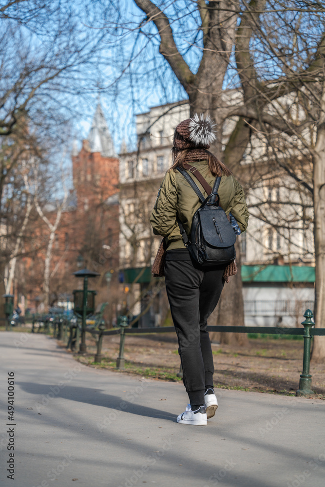 A lonely girl in a short jacket and sports trousers with a black bag behind her back walks along the asphalt in the city square on a sunny day in early spring. Back view