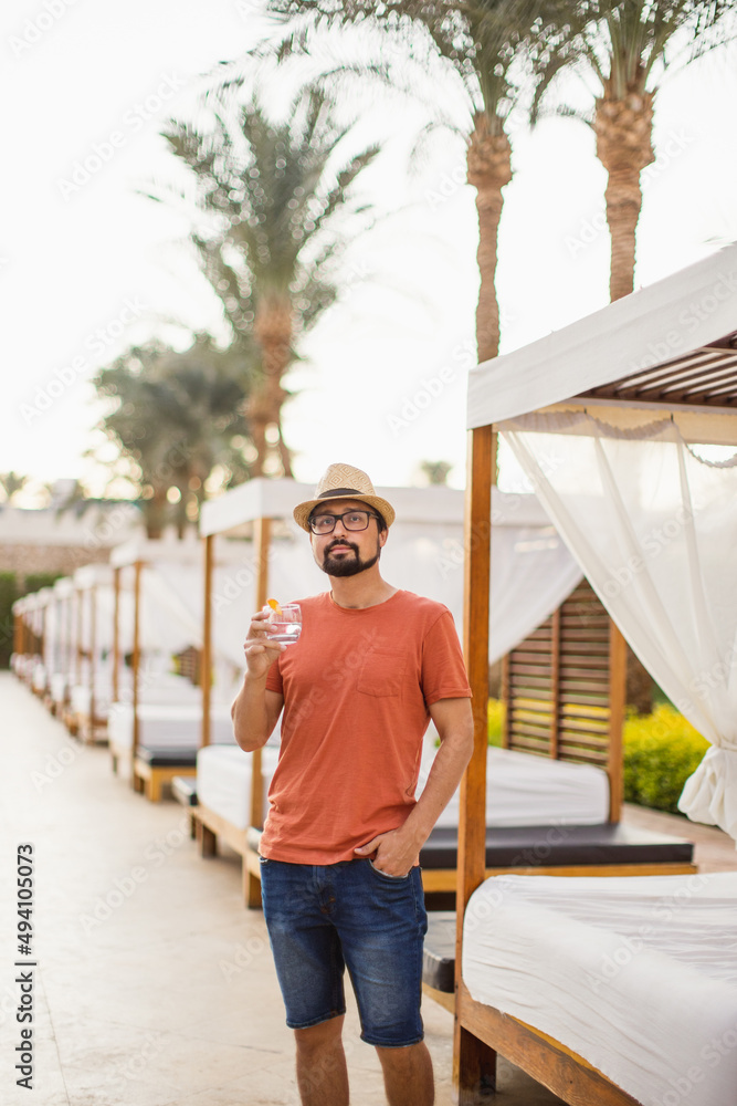 A young handsome man in an orange T-shirt, straw hat and denim shorts stands, drinking cool lemonade, looking away on the background of gazebos at a tropical luxury resort.