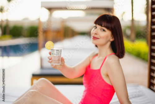 Close up of attractive female model in a pink swimsuit, lying in the beach gazebo bed and holding fresh drink, enjoying morning at luxury resort. Tropical paradise. Blur view of gazebo row