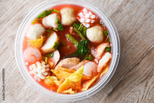 Red soup noodle pork balls crispy wontons with seafood squid shrimp and fish balls vegetable in soup bowl plastic on wooden table, top view Thai and China Asian food delivery concept