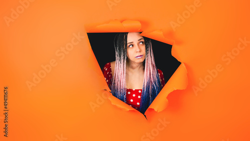 Young woman looking out of hole of orange background. Scared female looking around. © Anton Dios
