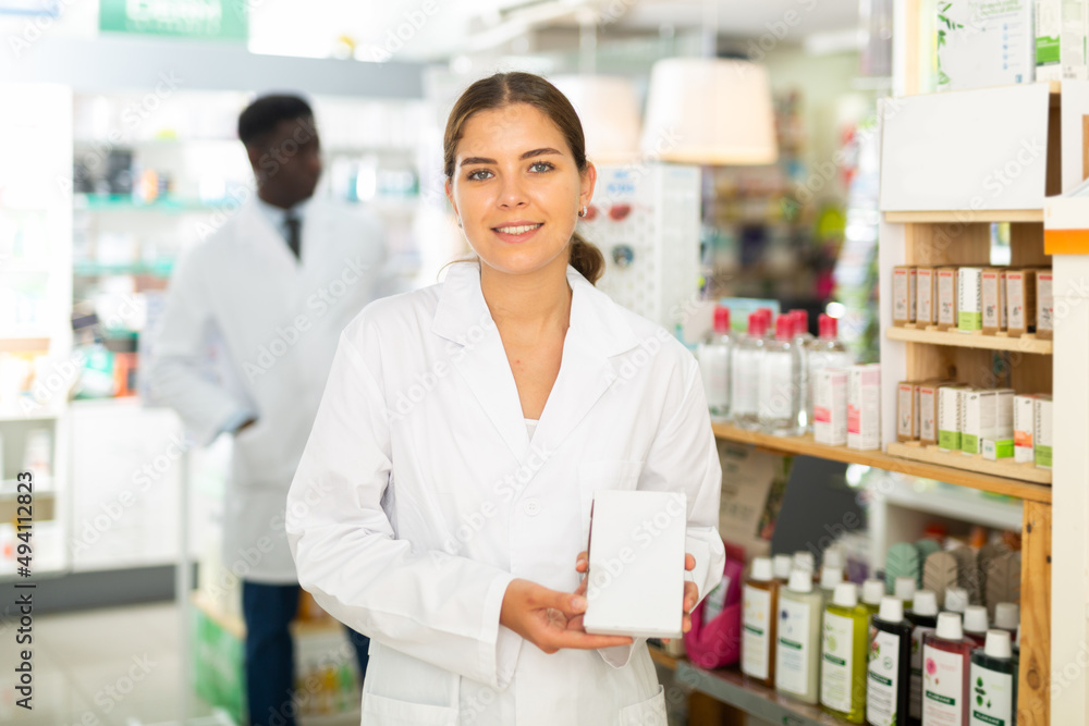 Female pharmacist standing with drug package in salesroom of drugstore. Her colleague standing behind.