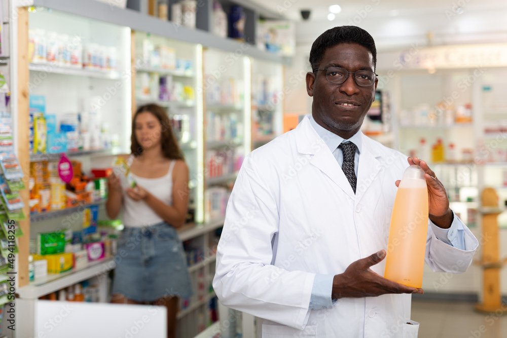 Male pharmacist standing in salesroom of drugstore and demonstrating haircare product. Client choosing drugs in background..