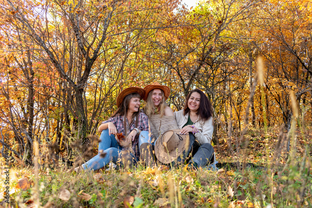 Three diverse female friends in cowboy hats are sitting on a log in the autumn forest. Three diverse female friends in cowboy hats are sitting on a log in the autumn forest.