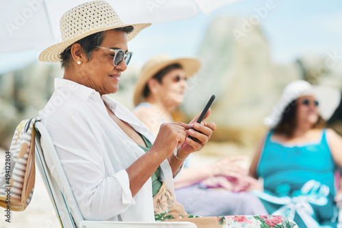 Sending all our selfies to our kids. Shot of a mature woman sitting alone and using her cellphone during a day out on the beach with friends.