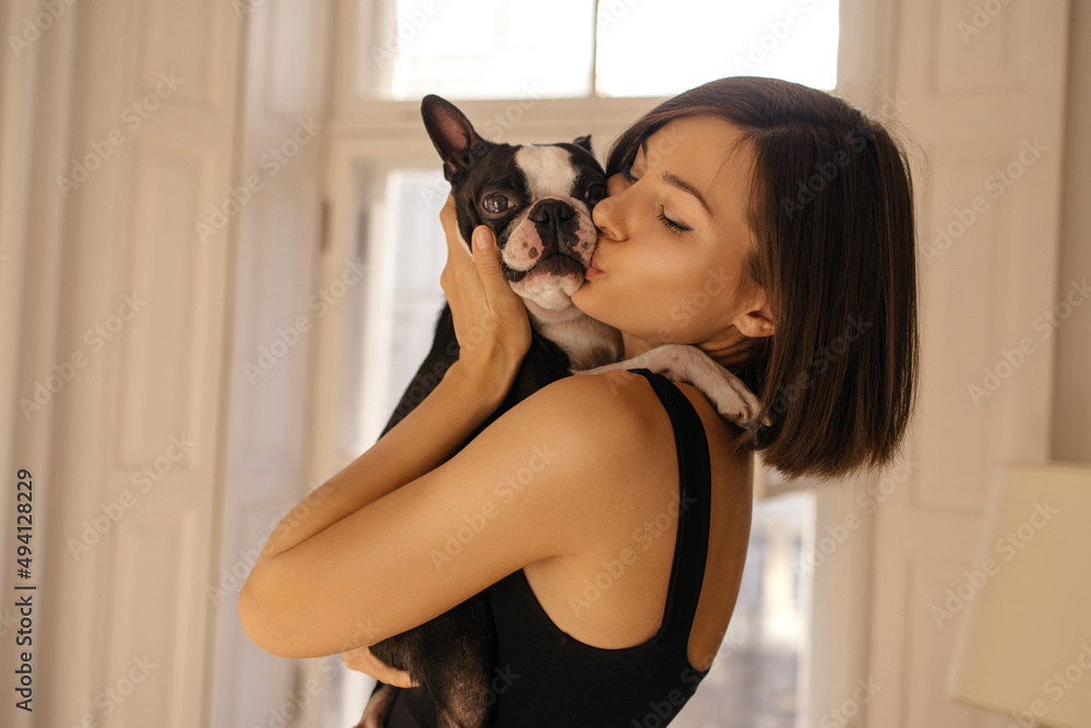 Cute young european woman kissing and hugging her boston terrier dog indoors. Brunette with bob haircut hugs and animal. Love between owner and pet