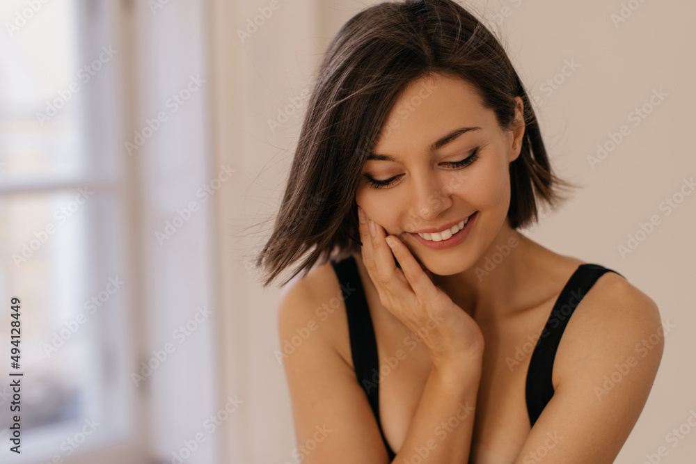 Close-up of young caucasian woman with beautiful smile looks down on white background. Dark-haired girl wears summer casual clothes. Concept of sensuality and femininity Close-up of young caucasian woman with beautiful smile looks down on white background. Dark-haired girl wears summer casual clothes. Concept of sensuality and femininity
