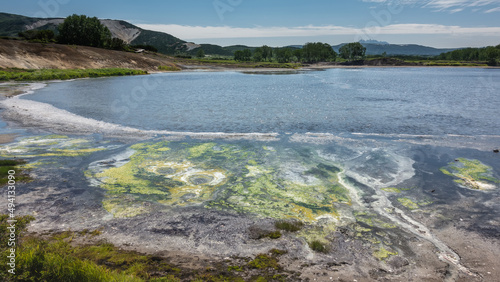 Turquoise lake in the caldera of the extinct volcano Uzon. Yellow-green clusters of thermophilic bacteria are visible off the coast. A mountain range against a blue sky. Kamchatka © Вера 