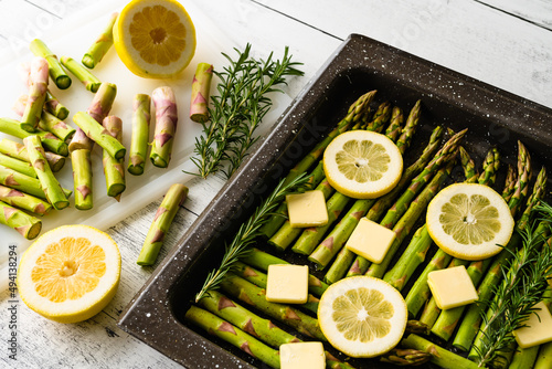 Fresh green asparagus, sliced lemon, and butter, fresh rosemary, and seasonig close up on baking sheet, ready to be baked, close-up view from above photo