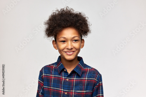 Laughing cheerful boy of 12 with stylish afro hair wearing plaid flannel shirt, isolated on white, looking aside, with cute lovely smile. Human emotions and feelings. Carefree childhood