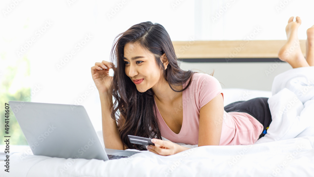 Asian young happy cheerful beautiful female model in casual pajama outfit lying down smiling hold credit card on bed in bedroom browsing surfing internet shopping online via laptop notebook computer Asian young happy cheerful beautiful female model in casual pajama outfit lying down smiling hold credit card on bed in bedroom browsing surfing internet shopping online via laptop notebook computer