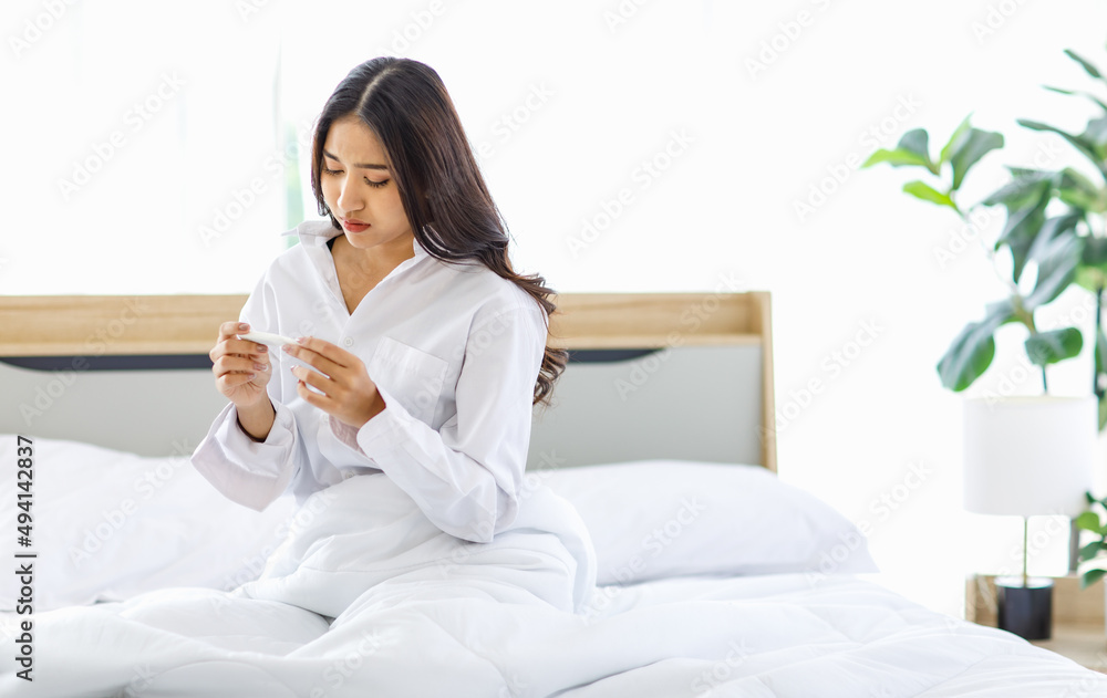 Asian young unhappy upset worried frowning face female teenager model wake up sitting under blanket on bed in bedroom holding pregnancy test kit and making phone call to boyfriend via smartphone Asian young unhappy upset worried frowning face female teenager model wake up sitting under blanket on bed in bedroom holding pregnancy test kit and making phone call to boyfriend via smartphone