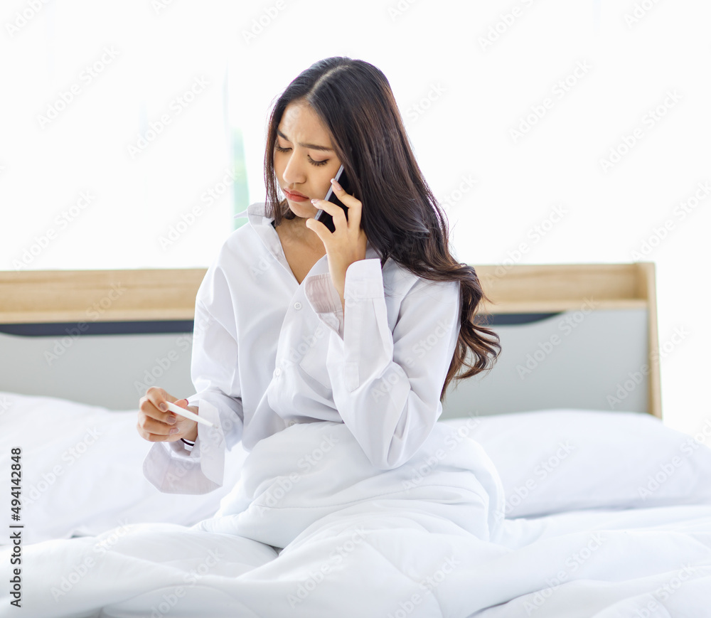 Asian young unhappy upset worried frowning face female teenager model wake up sitting under blanket on bed in bedroom holding pregnancy test kit and making phone call to boyfriend via smartphone Asian young unhappy upset worried frowning face female teenager model wake up sitting under blanket on bed in bedroom holding pregnancy test kit and making phone call to boyfriend via smartphone