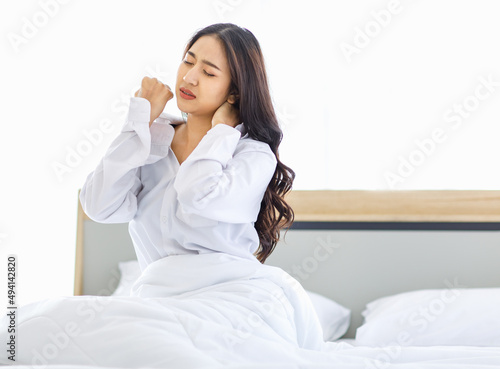Asian young unhappy upset frowning face female teenager model in casual pajama outfit waking up in morning sitting under white blanket on bed in bedroom holding hands on neck having painful ache