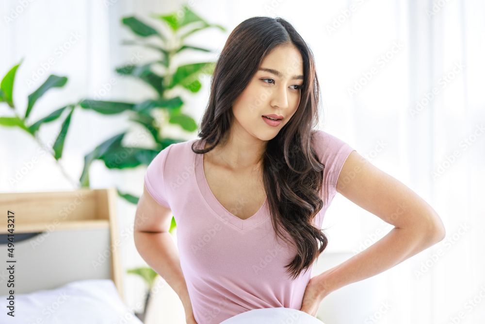 Asian young beautiful happy sleepy female teenager in casual pajama outfit waking up fresh in morning sitting smiling closed eyes under white blanket on bed in bedroom massaging stretching neck