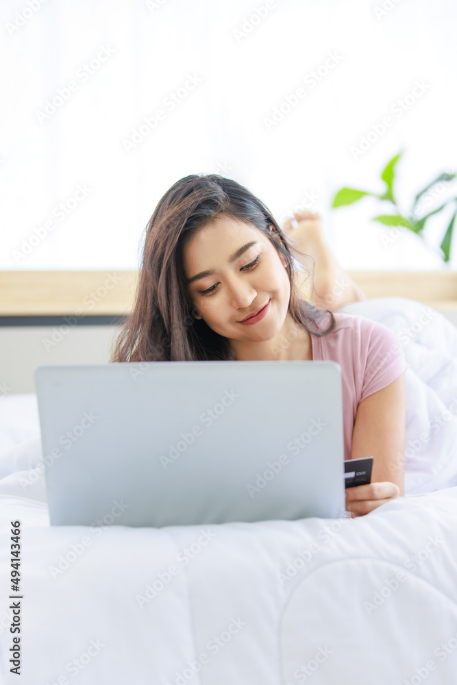 Asian young happy cheerful beautiful female model in casual pajama outfit lying down smiling hold credit card on bed in bedroom browsing surfing internet shopping online via laptop notebook computer Asian young happy cheerful beautiful female model in casual pajama outfit lying down smiling hold credit card on bed in bedroom browsing surfing internet shopping online via laptop notebook computer