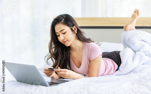 Asian young happy cheerful beautiful female model in casual pajama outfit lying down smiling hold credit card on bed in bedroom browsing surfing internet shopping online via laptop notebook computer