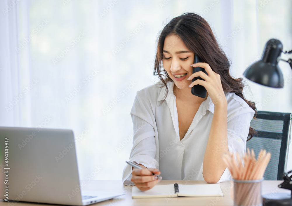 Asian young beautiful happy professional successful businesswoman designer sitting smiling at workstation desk using laptop notebook computer and smartphone working remotely online at home office Asian young beautiful happy professional successful businesswoman designer sitting smiling at workstation desk using laptop notebook computer and smartphone working remotely online at home office