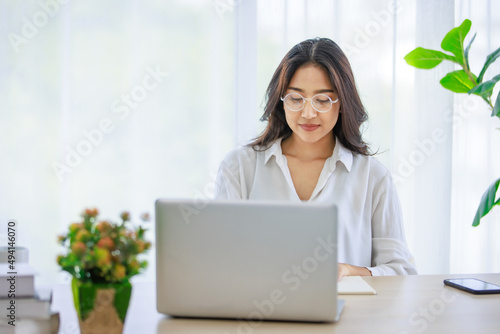 Asian young beautiful female university student wearing optical eyeglasses sitting at working desk remotely studying learning streaming online via laptop notebook computer at home during quarantine © Bangkok Click Studio