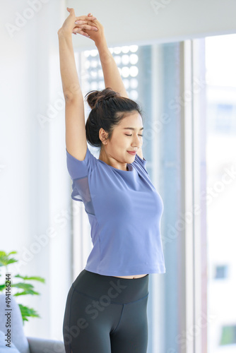 Asian young beautiful happy healthy female sport girl in casual sporty outfit standing smiling holding hand up stretching arm and body exercising training routine yoga pilates practice in living room © Bangkok Click Studio