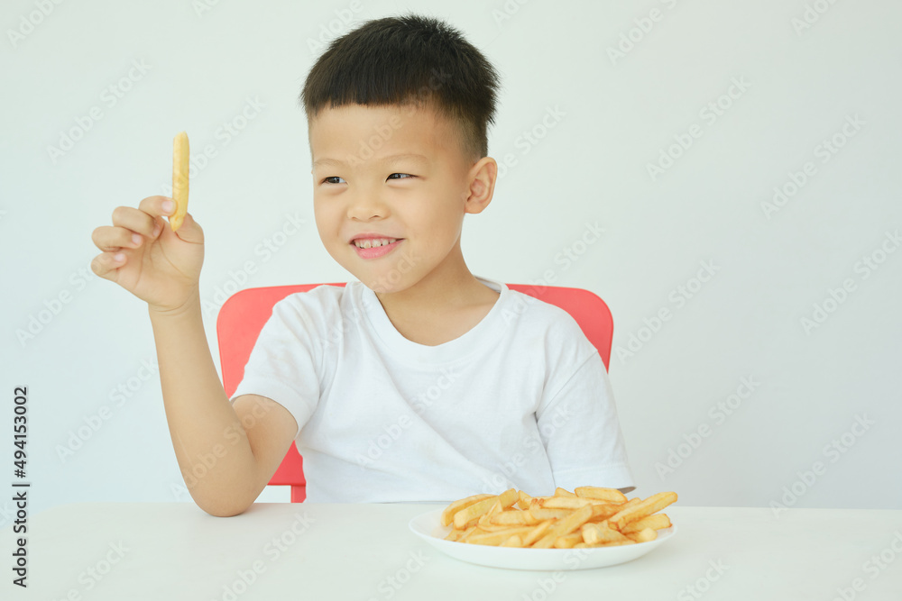 Cute happy smiling Asian 5 yers old kindergarten boy child eating French fries potato chips sitting at the table Isolated over white wall background, Unhealthy Kids Foods concept Cute happy smiling Asian 5 yers old kindergarten boy child eating French fries potato chips sitting at the table Isolated over white wall background, Unhealthy Kids Foods concept