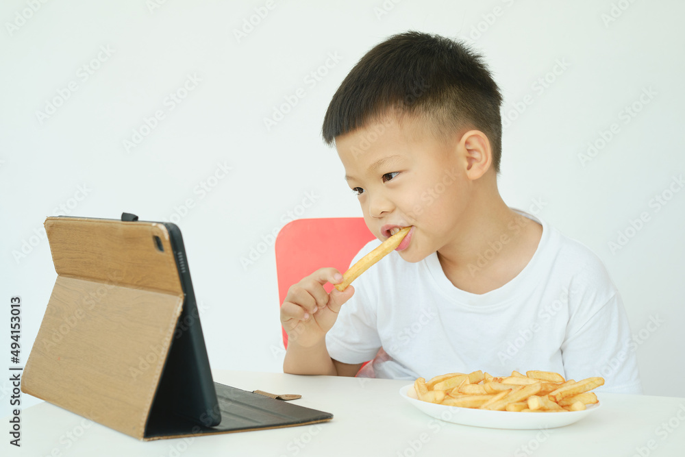 Asian 5 years old little kindergarten boy child eating French fries while watching a video from tablet pc isolated over white wall background, Unhealthy Kids Foods, Gadget-addicted children concept Asian 5 years old little kindergarten boy child eating French fries while watching a video from tablet pc isolated over white wall background, Unhealthy Kids Foods, Gadget-addicted children concept