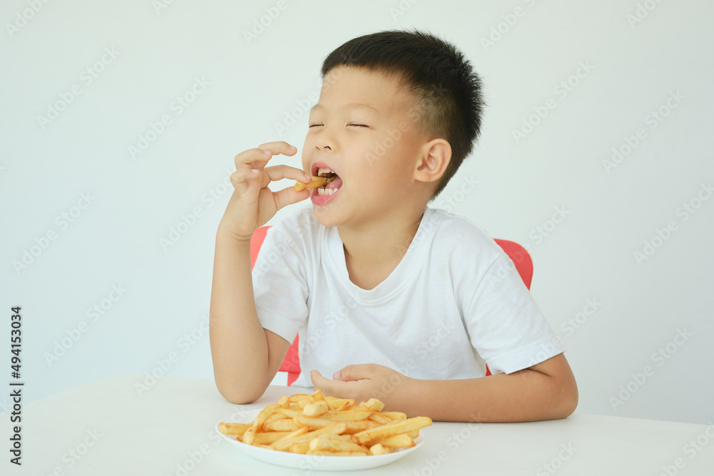 Cute happy smiling Asian 5 yers old kindergarten boy child eating French fries potato chips sitting at the table Isolated over white wall background, Unhealthy Kids Foods concept Cute happy smiling Asian 5 yers old kindergarten boy child eating French fries potato chips sitting at the table Isolated over white wall background, Unhealthy Kids Foods concept