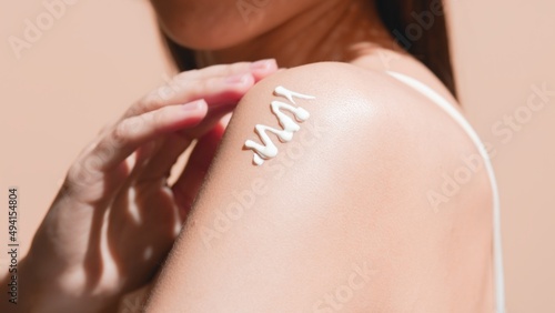 Young slim woman with long dark hair applies sunblock on her shoulder with her hand against beige background | Sunblock applying shot for body care commercial photo