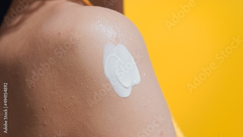Young slim dark-haired woman in a yellow swimwear applies sun cream on her shoulder against yellow background | Sun cream application shot for body care commercial