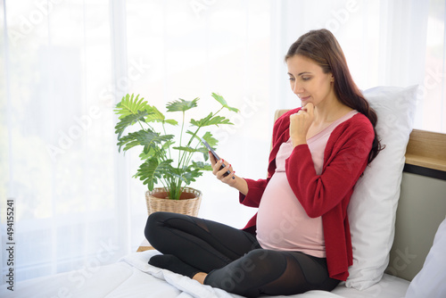 Pregnant woman sitting on bed and using smart phone in modern bedroom.