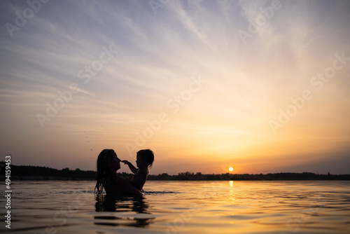 Mom plays with a naked baby in oversleeves in the lake against the background of a summer sunset