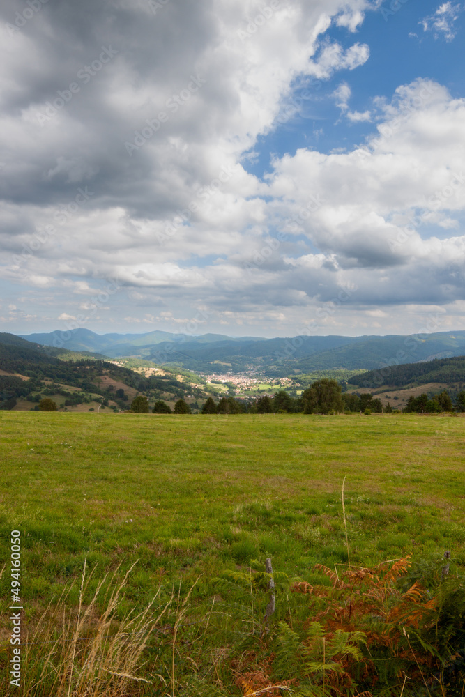 Vosges mountain landscape in the summer. The sky is overcast and a little blue. A large green meadow slopes down to the valley. In the distance another massif stands out. Vosges mountain landscape in the summer. The sky is overcast and a little blue. A large green meadow slopes down to the valley. In the distance another massif stands out.