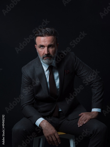 Portrait of adult businessman wearing trendy suit and sitting in modern studio on stylish chair against the black background