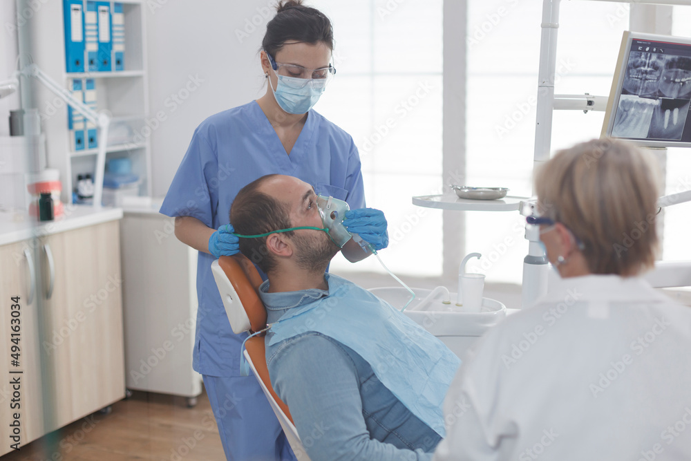 Man patient sitting on dental chair during stomatology examination while dentist assistant putting oxygen mask before teeth surgery in stomatological office room. Medical team wearing face mask Man patient sitting on dental chair during stomatology examination while dentist assistant putting oxygen mask before teeth surgery in stomatological office room. Medical team wearing face mask