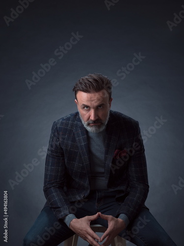 Portrait of adult businessman wearing trendy suit and sitting in modern studio on stylish chair against the black background