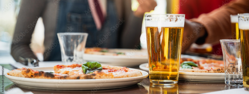 Vertical Close-up glass of beer with froth on table next to a plate with Margherita pizza. Unrecognizable group of people unfocused on background enjoy and eating