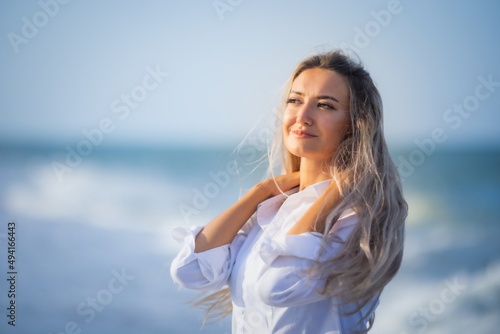 A girl with long hair in a bluish swimsuit and a shirt goes through her hair on the seashore