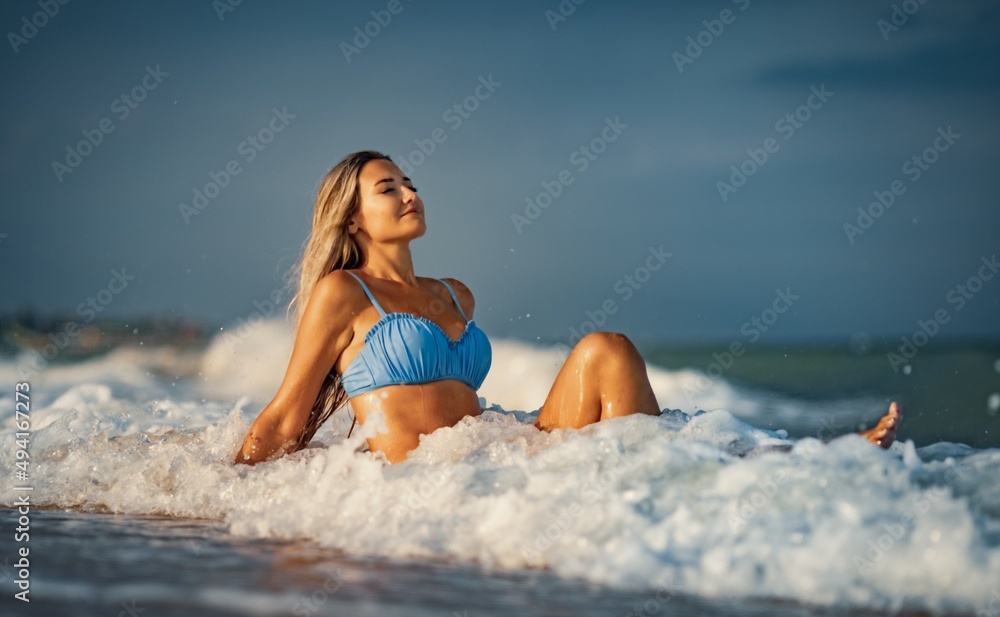A girl with blond hair in a delicate swimsuit sits by the sea, enjoying the splash of waves A girl with blond hair in a delicate swimsuit sits by the sea, enjoying the splash of waves